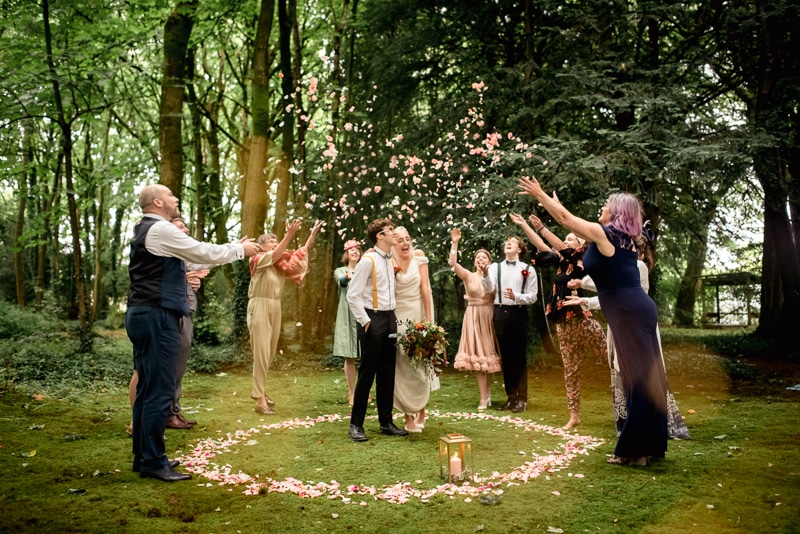 A bride and groom stand int he centre of a petal circle in a woodland. The ground is green and mossy. The celebrant and guests throw petals into the air over the heads of the couple