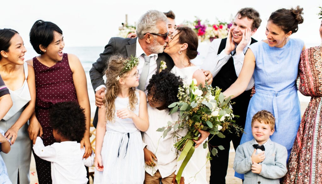Informal wedding shot. There are children standing in front of a man and woman who are kissing. The groom has silver hair and a beard and wears a grey suit. The bride appears younger , has long dark hair in a bun, a white dress and holds a large bunch of flowers. There are happy looking people around the couple