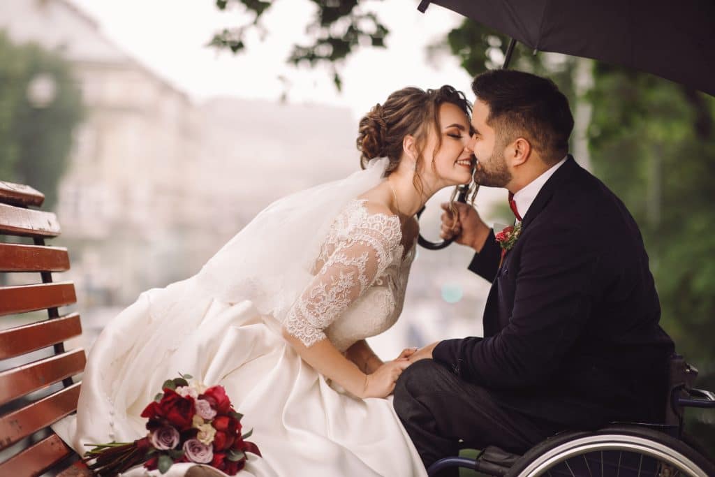 Seated bride in white wedding dress leans  over to kiss handsome groom in black suit. Groom is in a wheelchair.