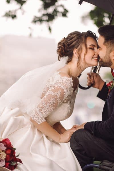 Seated bride in white wedding dress leans over to kiss handsome groom in black suit. Groom is in a wheelchair.